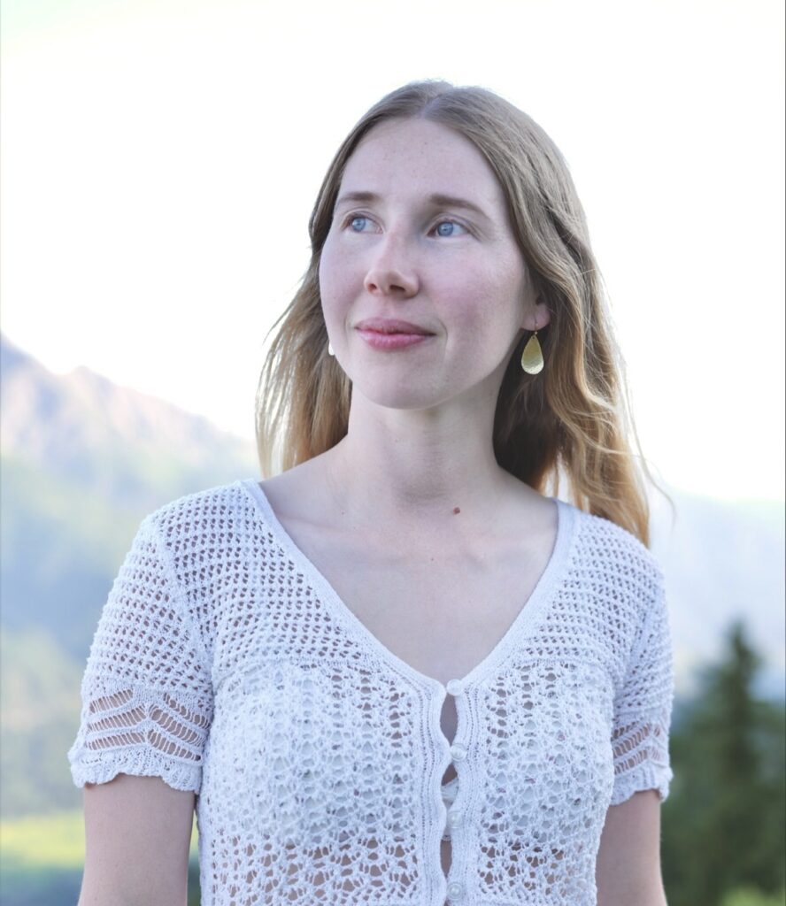 Clara Tiefenthaler in einem weissen Häkeloberteil, sie blickt lächelnd zur Seite, dahinter unscharfe Berge.